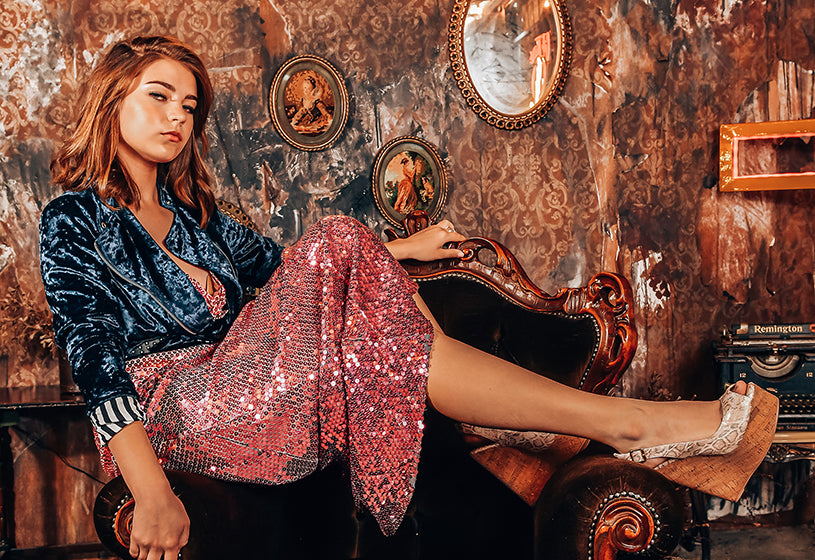 Female model wears a blue velvet jacket, pink sequin gown dress and pink snake print platform wedge shoes. She is sitting over a vintage velvet armchair.